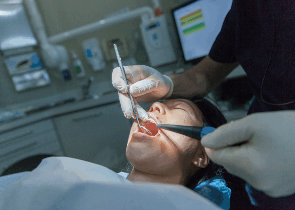 A woman sits in a dental chair while a dentist cleans her teeth with dental tools.